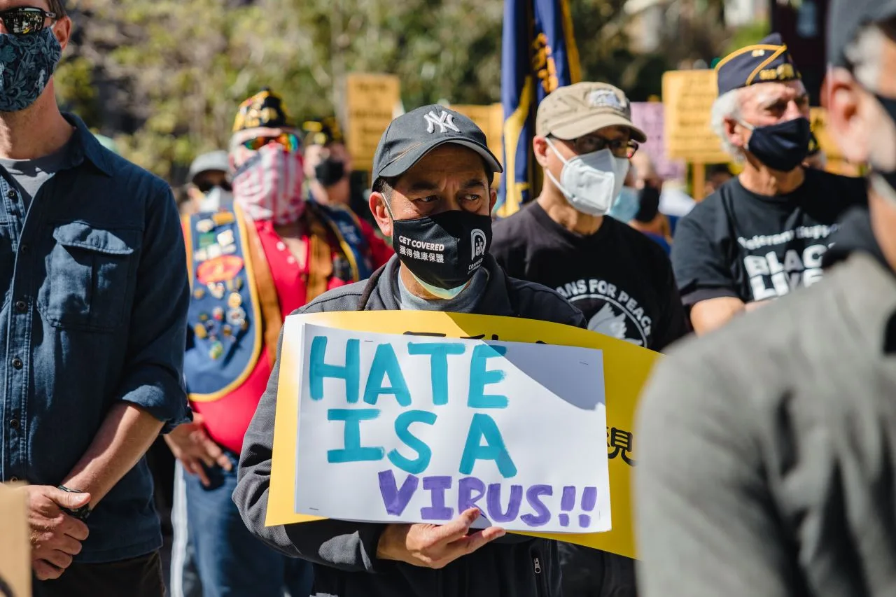 Masked man holding a sign that says "HATE IS A VIRUS!!" in a rally