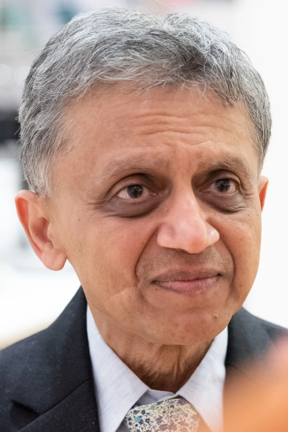 A close portrait in a dark suit, pale shirt, and a patterned tie, grey hair swept back, looking just past the camera.