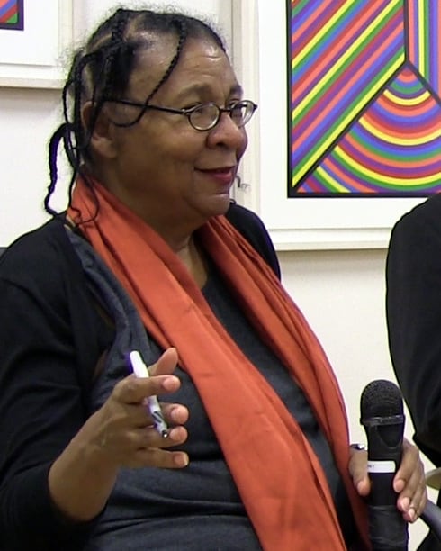 Speaking at a seated public event in a black cardigan and a wide orange scarf, a microphone held beside her.