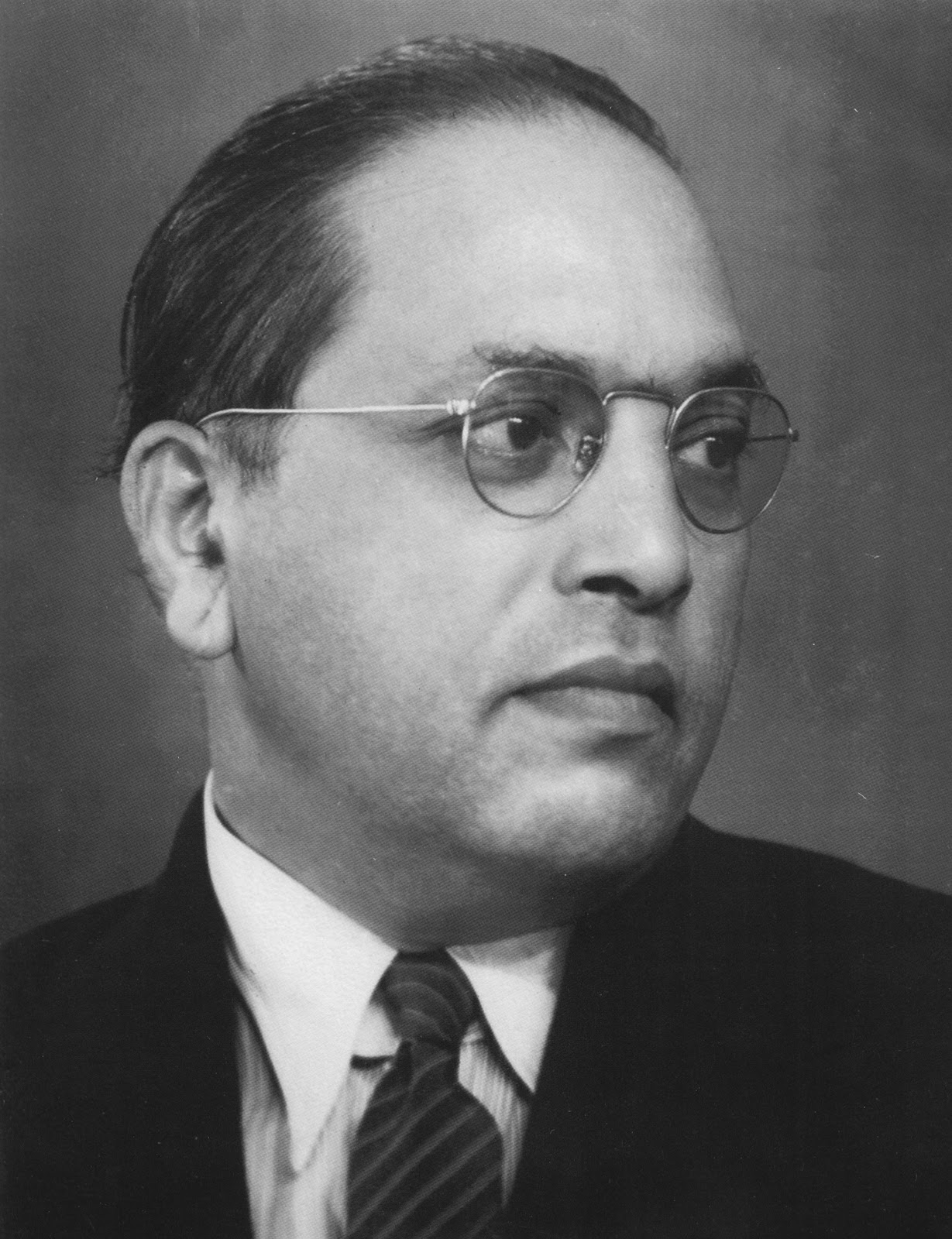 A studio portrait in three-quarter profile, in a dark suit, striped tie, and round wire-rimmed glasses.