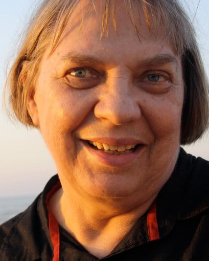 Smiling into the camera on a beach at sunset, in a dark shirt, the sea behind her.